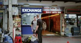 Two men with backpacks stand outside a currency exchange in Bangkok, Thailand.