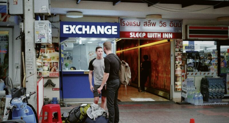 Two men with backpacks stand outside a currency exchange in Bangkok, Thailand.